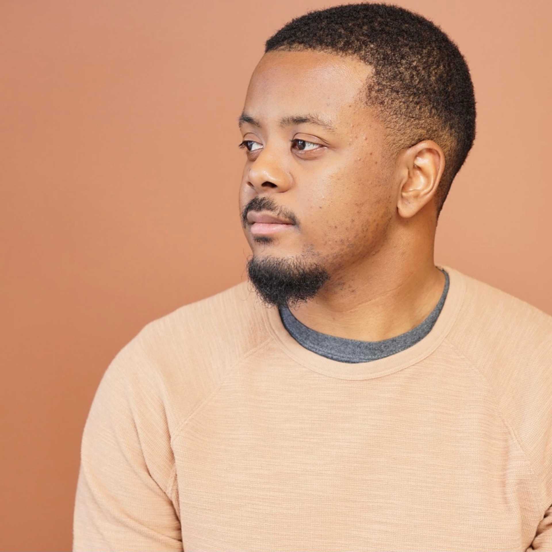 Man in beige sweater looking thoughtfully to the side against a brown background.