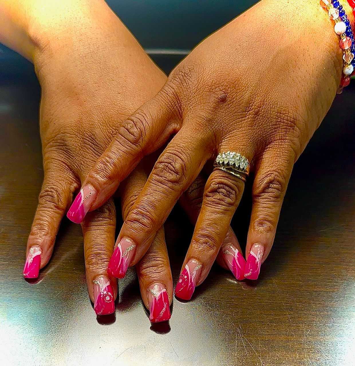 Hands with pink, glittery manicured nails and a diamond ring on finger, resting on a wooden surface.