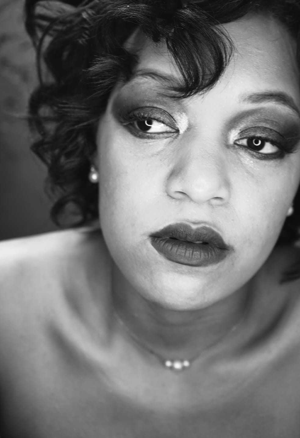 Black and white portrait of a woman with curly hair, wearing makeup and a delicate necklace.