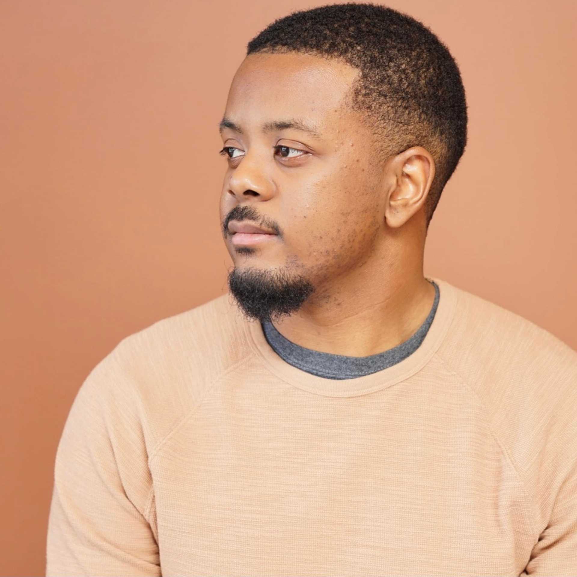 Man in beige sweater looking thoughtfully to the side against a brown background.